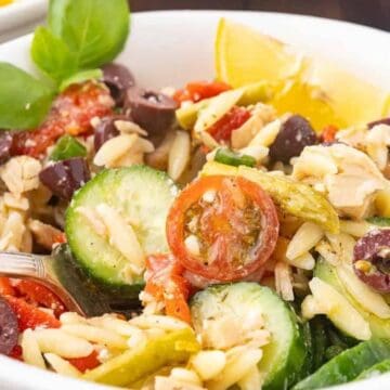 A white bowl filled with orzo pasta salad, featuring sliced cucumbers, cherry tomatoes, black olives, green beans, red peppers, and chunks of tuna, garnished with a lemon wedge and fresh basil leaves. A fork is in the bowl.