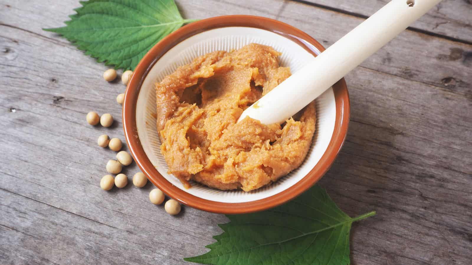 A bowl of brown miso paste with a white pestle resting inside, surrounded by soybeans and green leaves on a wooden surface.