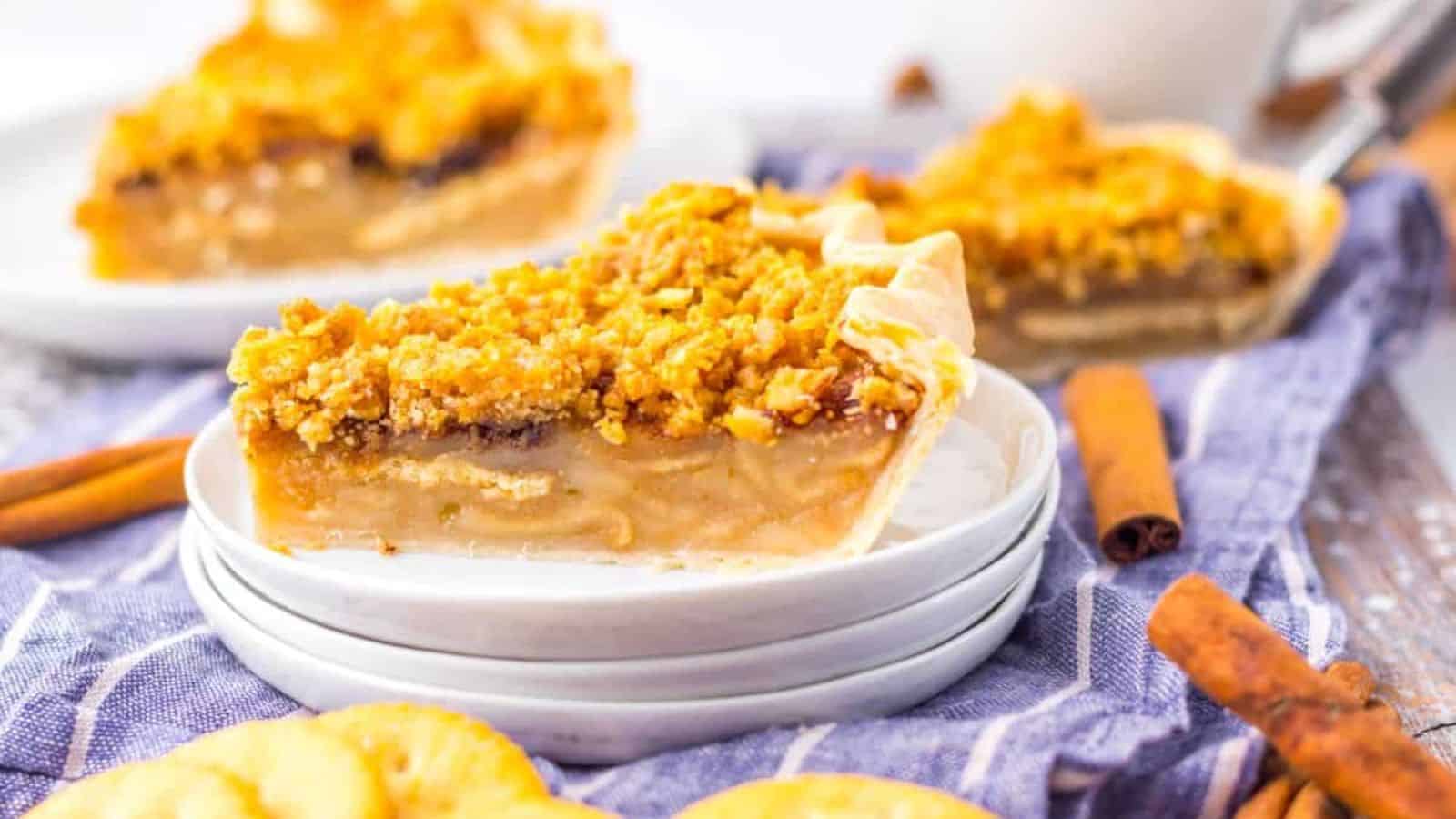 Slice of pie with crumb topping on a stack of plates, surrounded by crackers and cinnamon sticks.