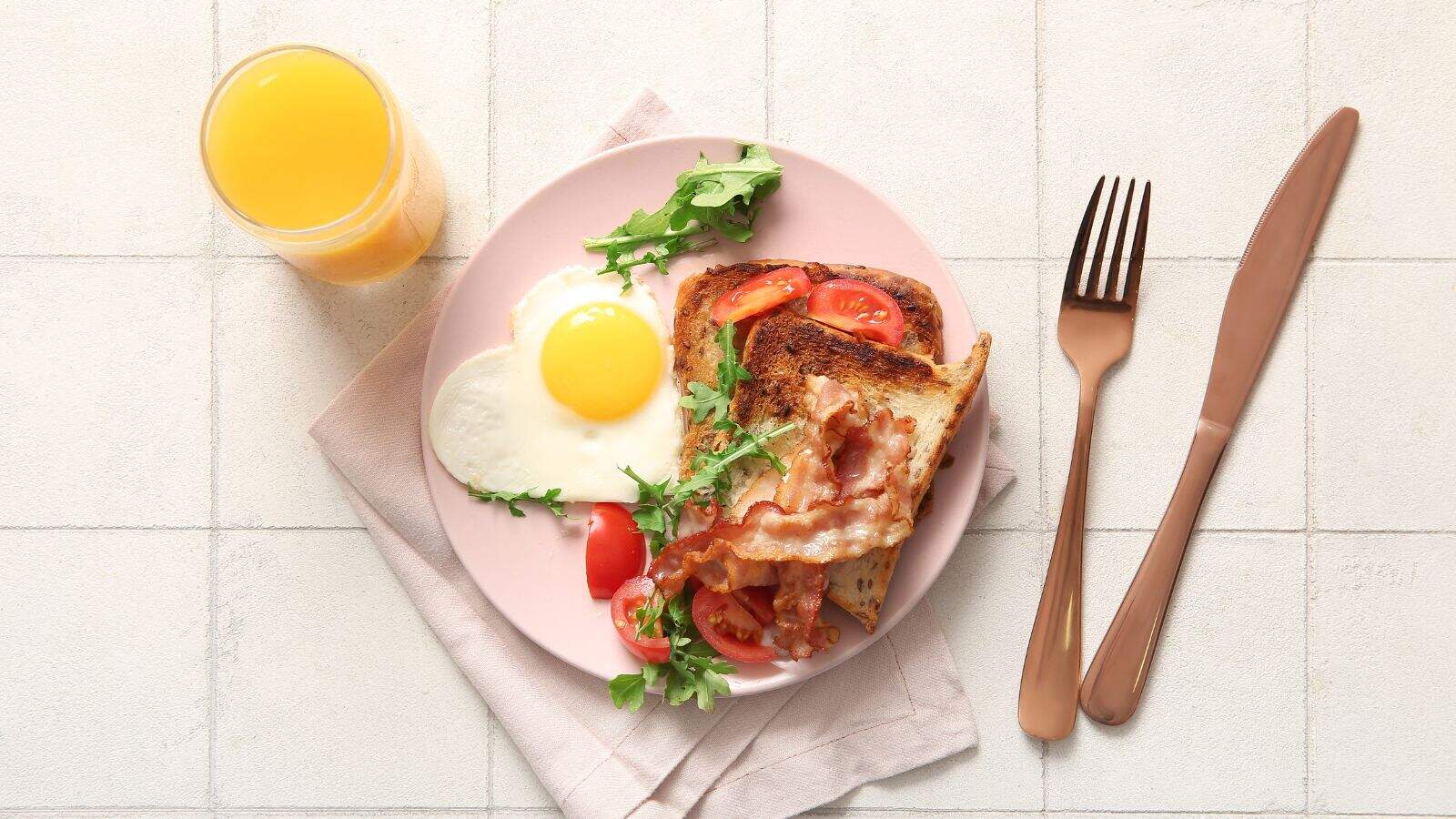 A pink plate with toast topped with bacon, tomato, and greens, a fried egg, and a few cherry tomatoes, next to a glass of orange juice. A fork and knife are placed on the right, on a light colored surface.