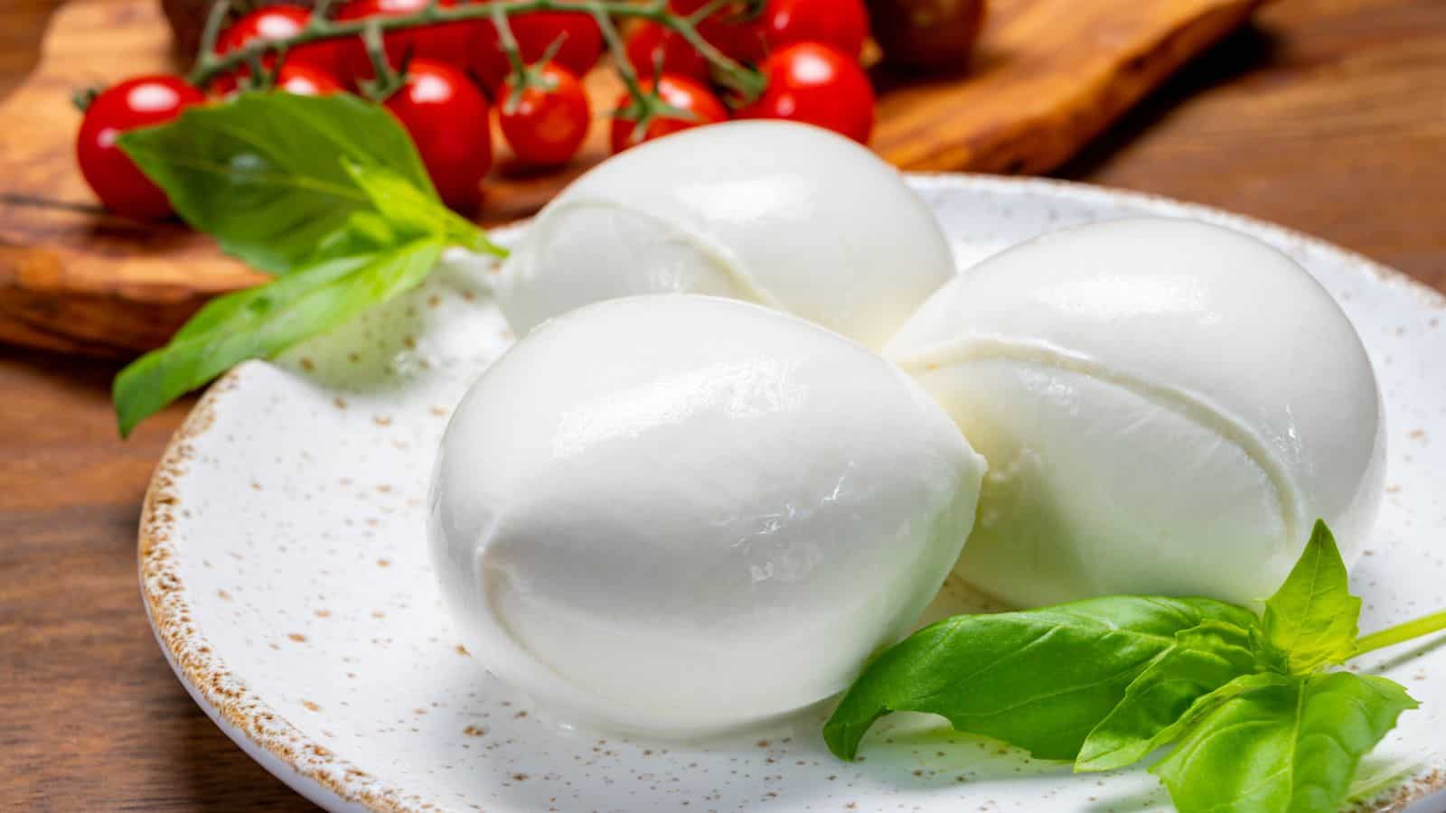 Three balls of fresh mozzarella cheese sit on a white speckled plate, garnished with basil leaves. In the background, there is a wooden board with a cluster of cherry tomatoes.