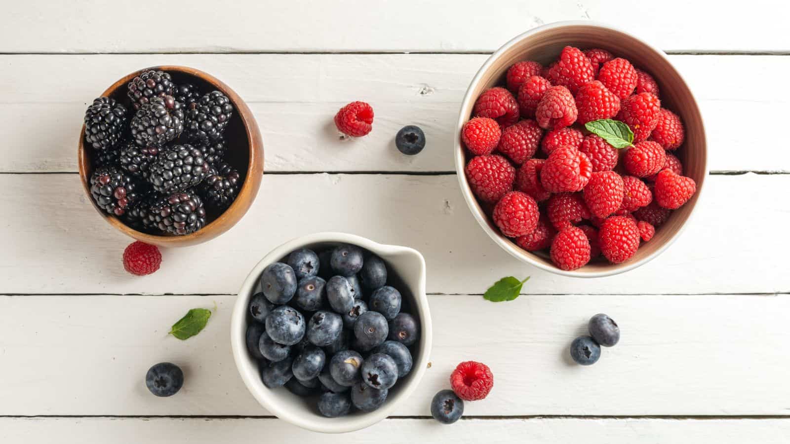 Three bowls of fresh berries on a white wooden surface: one with blackberries, one with raspberries garnished with a mint leaf, and one with blueberries. A few loose berries and mint leaves are scattered around.