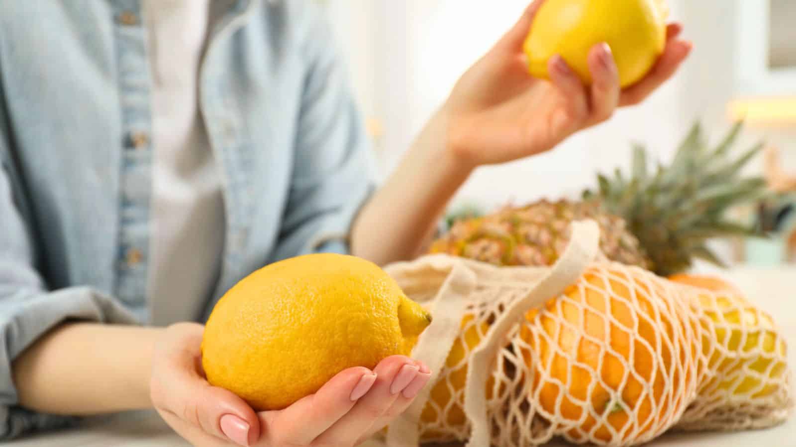A person holding two lemons, with a reusable mesh bag containing a pineapple and other fruits in the background. Only the person's hands and part of their upper body are visible.