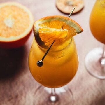 Two glasses of orange-colored cocktails garnished with orange slices and skewers are on a wooden surface. A halved orange is also visible in the background.