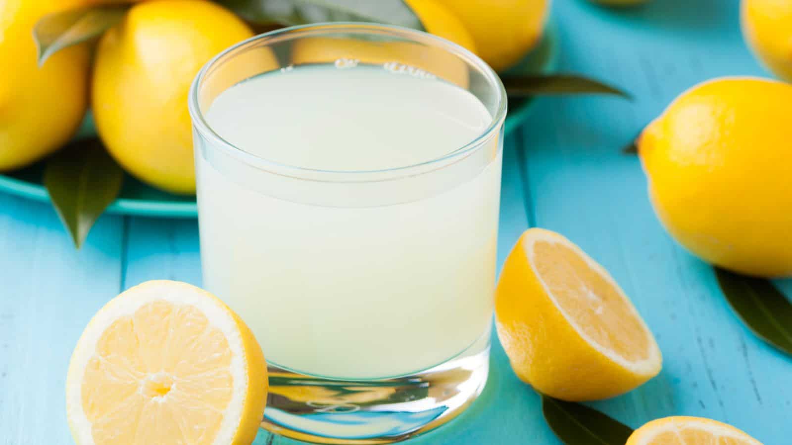 A clear glass filled with lemon juice is placed on a blue wooden surface, surrounded by whole lemons and a halved lemon, some with attached green leaves. A plate with more lemons is visible in the background.