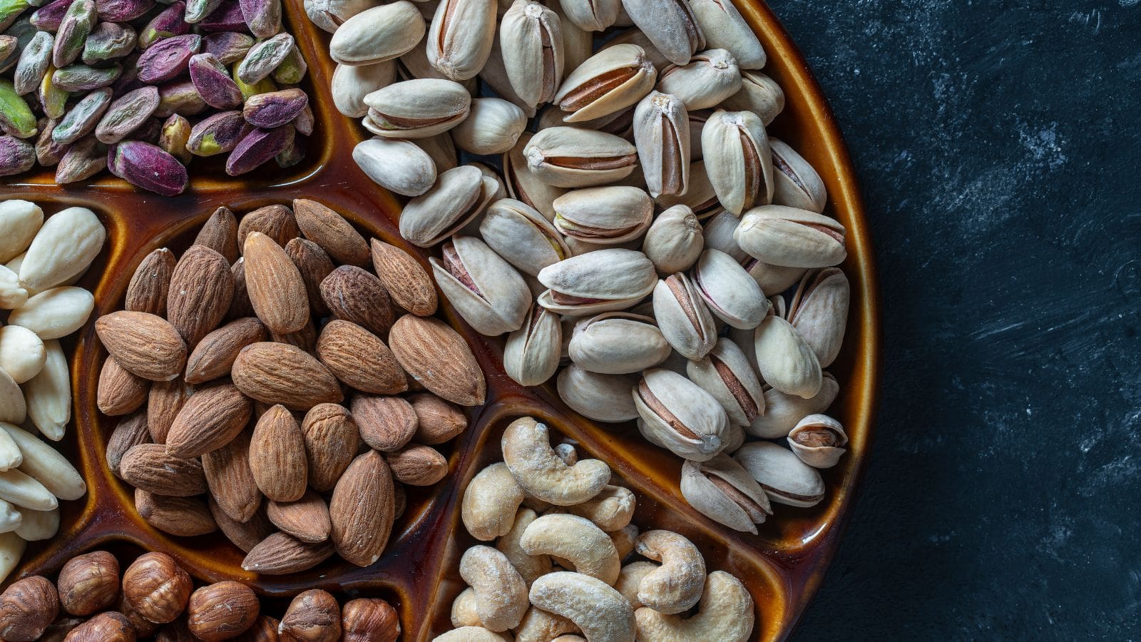 A close-up photo of a round tray divided into sections, each containing a different type of nut, including pistachios, almonds, cashews, hazelnuts, and peeled almonds, on a dark textured background.