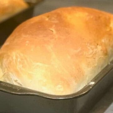 A loaf of golden-brown bread sits in a rectangular metal baking pan on a countertop, with another loaf partially visible in the background.