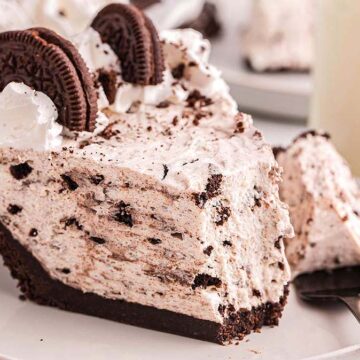 A close-up of a slice of cookies and cream pie with a chocolate cookie crust, topped with whipped cream and whole chocolate sandwich cookies, on a white plate with a fork. A glass of milk is in the background.