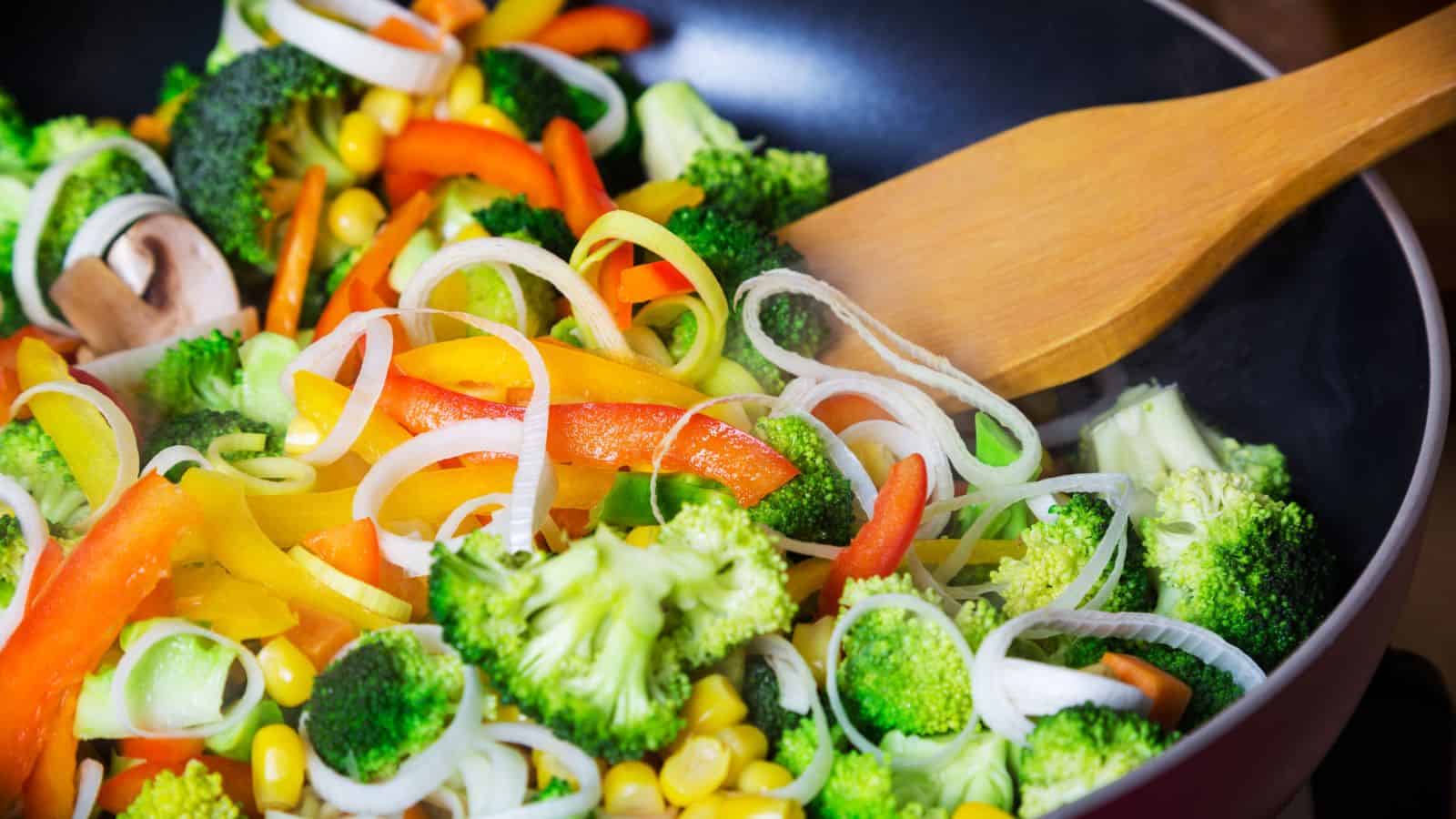 A close-up of a pan with assorted vegetables including broccoli, sliced onions, red and yellow bell peppers, corn, and mushrooms being stirred with a wooden spatula.