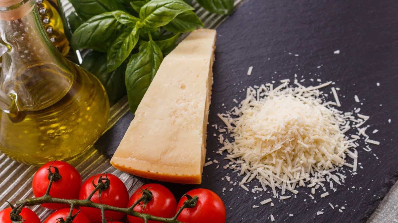 A wedge of parmesan cheese, a small pile of grated parmesan, a bunch of fresh basil leaves, a bottle of olive oil, and several vine tomatoes are arranged on a dark slate surface.