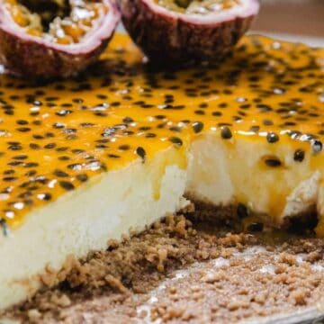 A cheesecake topped with a glossy layer of passion fruit and seeds, with a crumbly biscuit base. A slice has been cut and removed, and two halved passion fruits rest on top of the cheesecake.