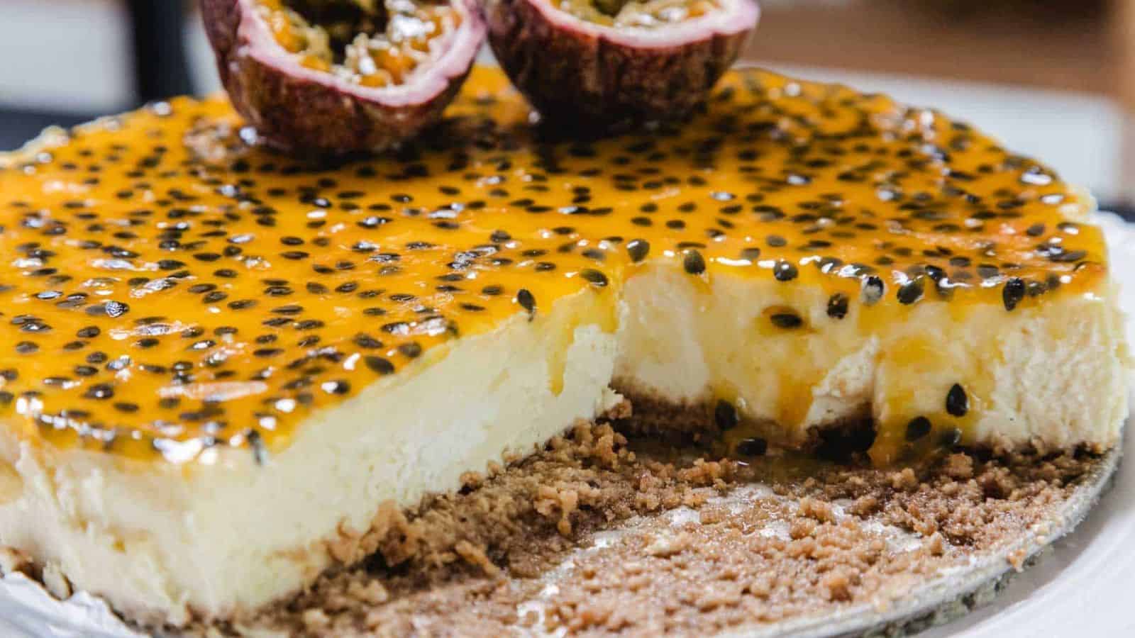 A cheesecake topped with a glossy layer of passion fruit and seeds, with a crumbly biscuit base. A slice has been cut and removed, and two halved passion fruits rest on top of the cheesecake.