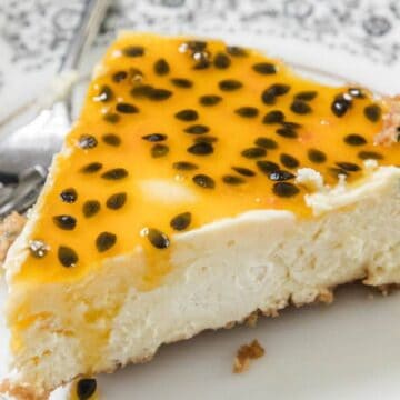 A slice of cheesecake topped with a glossy yellow passionfruit sauce and black seeds sits on a decorative white plate with a fork beside it.