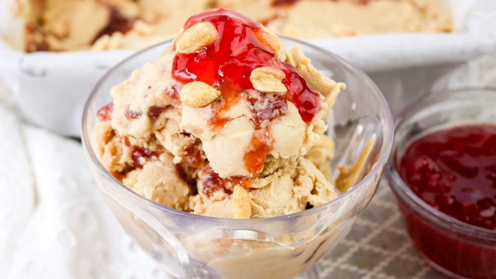 A glass bowl of peanut butter ice cream topped with red jelly and peanuts. A container and a bowl of red jelly are in the background.