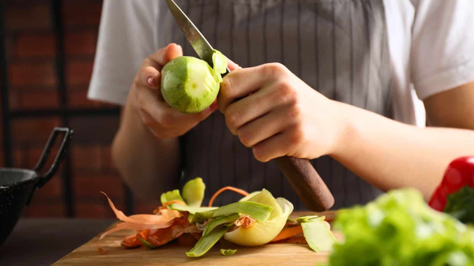 A person wearing a gray apron uses a knife to peel a round green vegetable on a wooden cutting board, with onion skins and other vegetable scraps nearby.