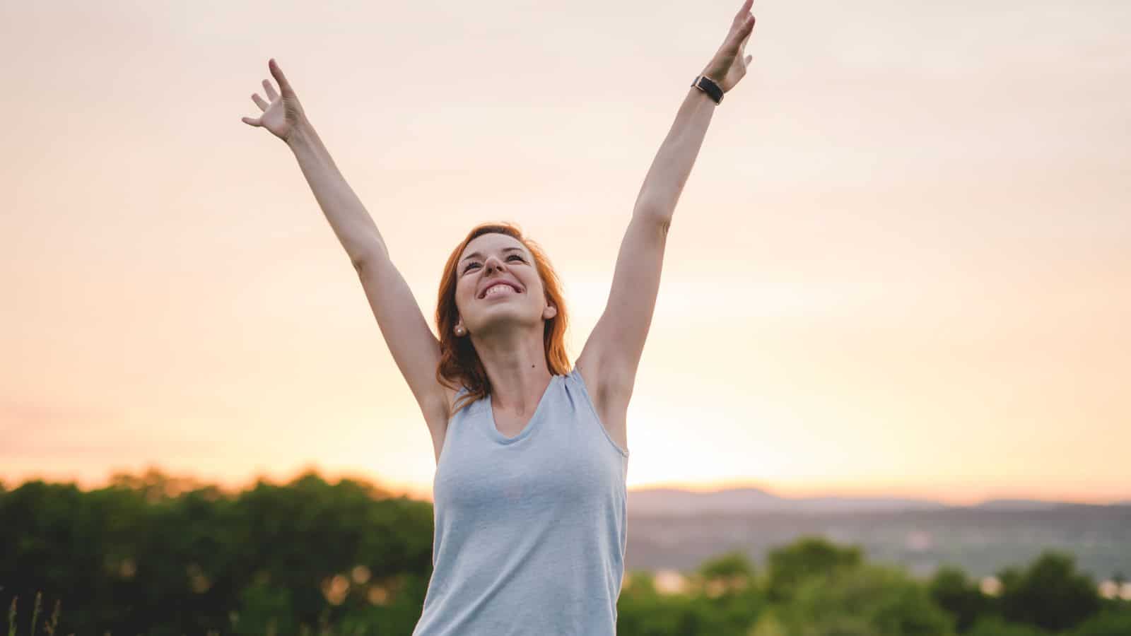A person stands outdoors with arms raised and smiling, wearing a sleeveless top. The sky is bright with a warm glow, and trees and hills are visible in the background.
