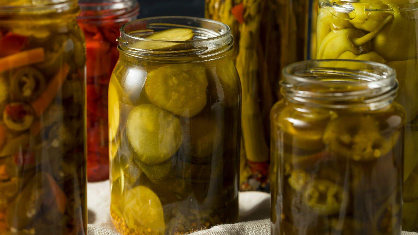 Several glass jars filled with assorted pickled vegetables, including cucumber slices and peppers, are arranged closely together on a surface. The contents are visible through the clear jars.