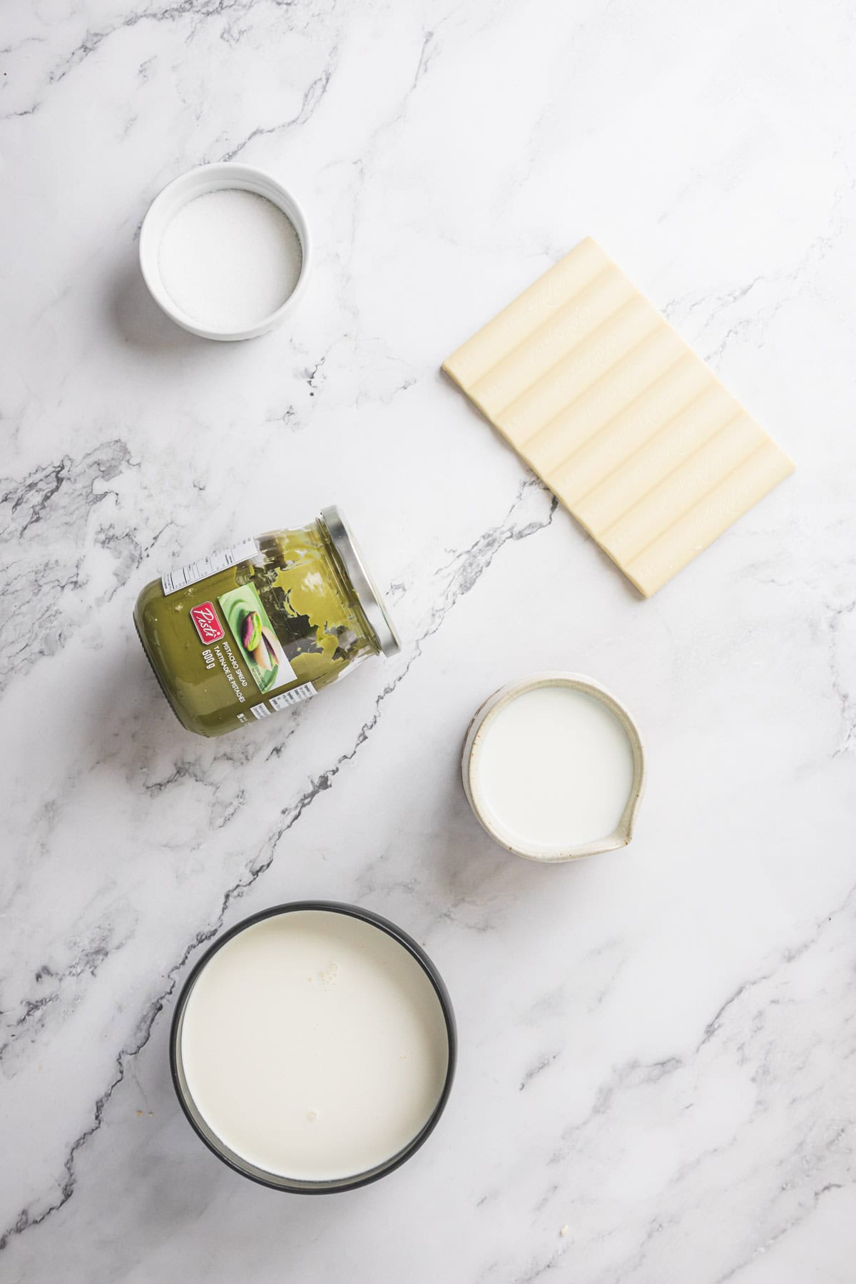 A flat lay of lasagna ingredients on a marble surface, featuring a bowl of milk, a small jug of cream, a jar of pickles, a small bowl of salt, several sheets of uncooked lasagna noodles, and a scoop of pistachio ice cream for dessert.