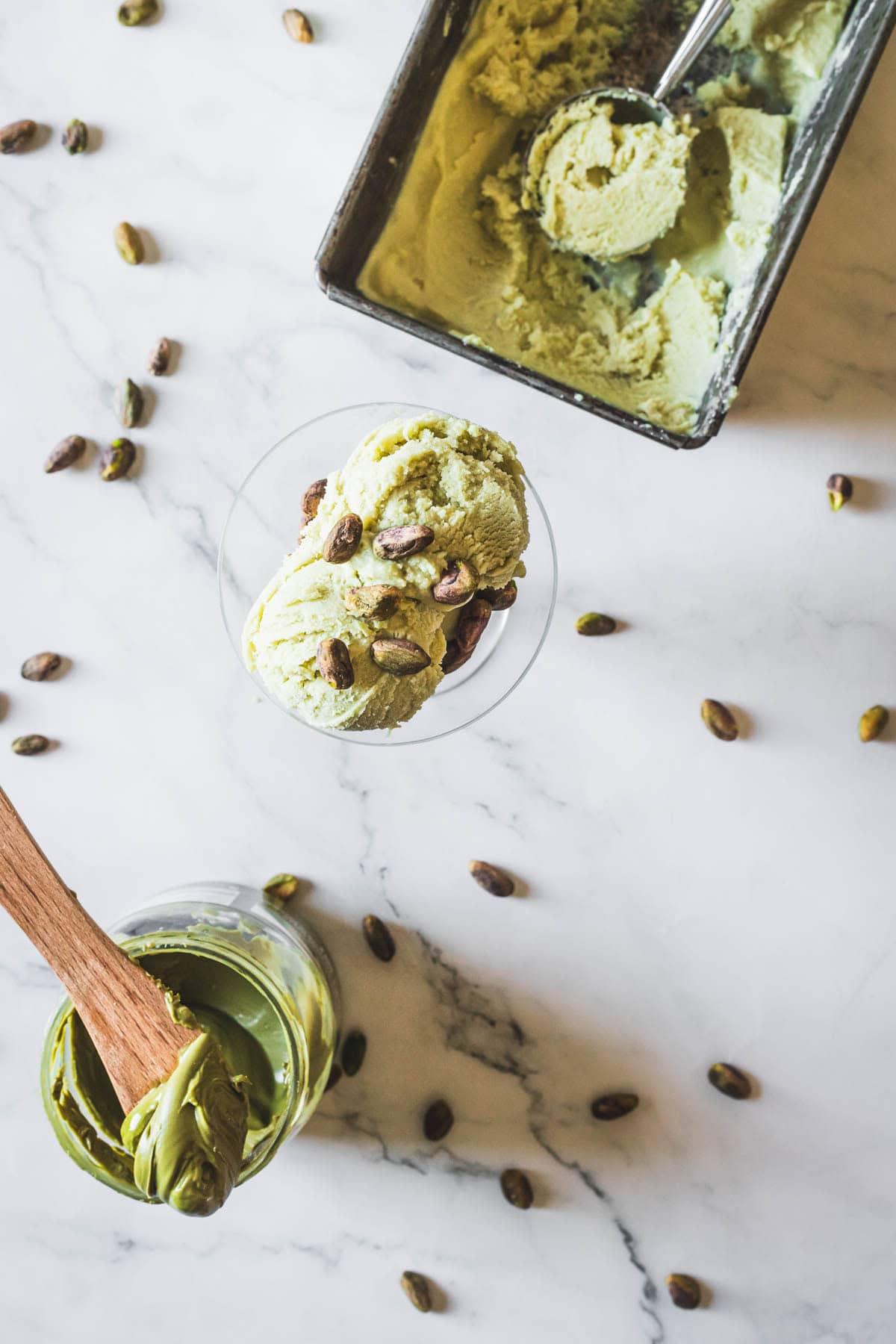 Overhead view of Pistachio Ice Cream scoops in a glass, topped with pistachios. A container of Pistachio Ice Cream with a scoop, scattered pistachios, and a jar with a wooden spoon rest on a marble surface.