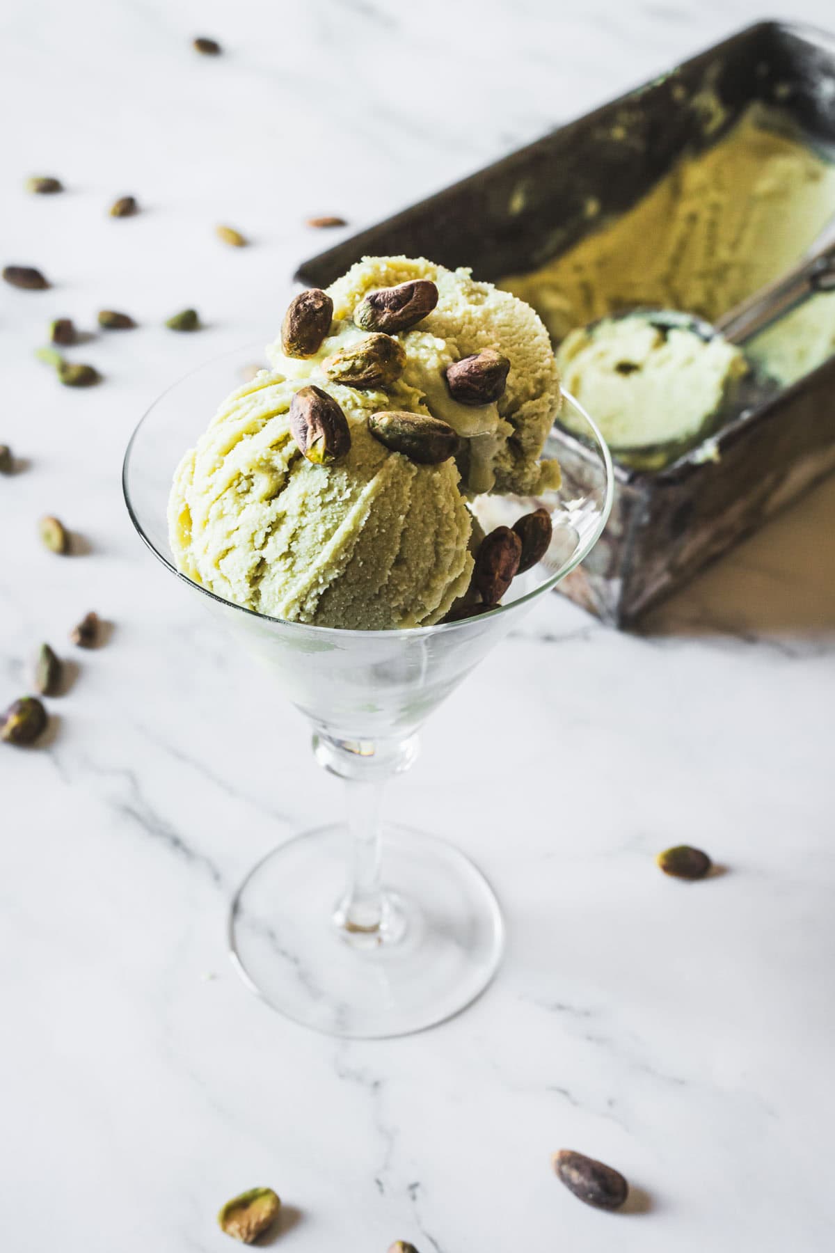 A glass holds three scoops of creamy Pistachio Ice Cream topped with whole pistachios, with extra pistachios scattered on a marble surface and a metal ice cream container in the background.