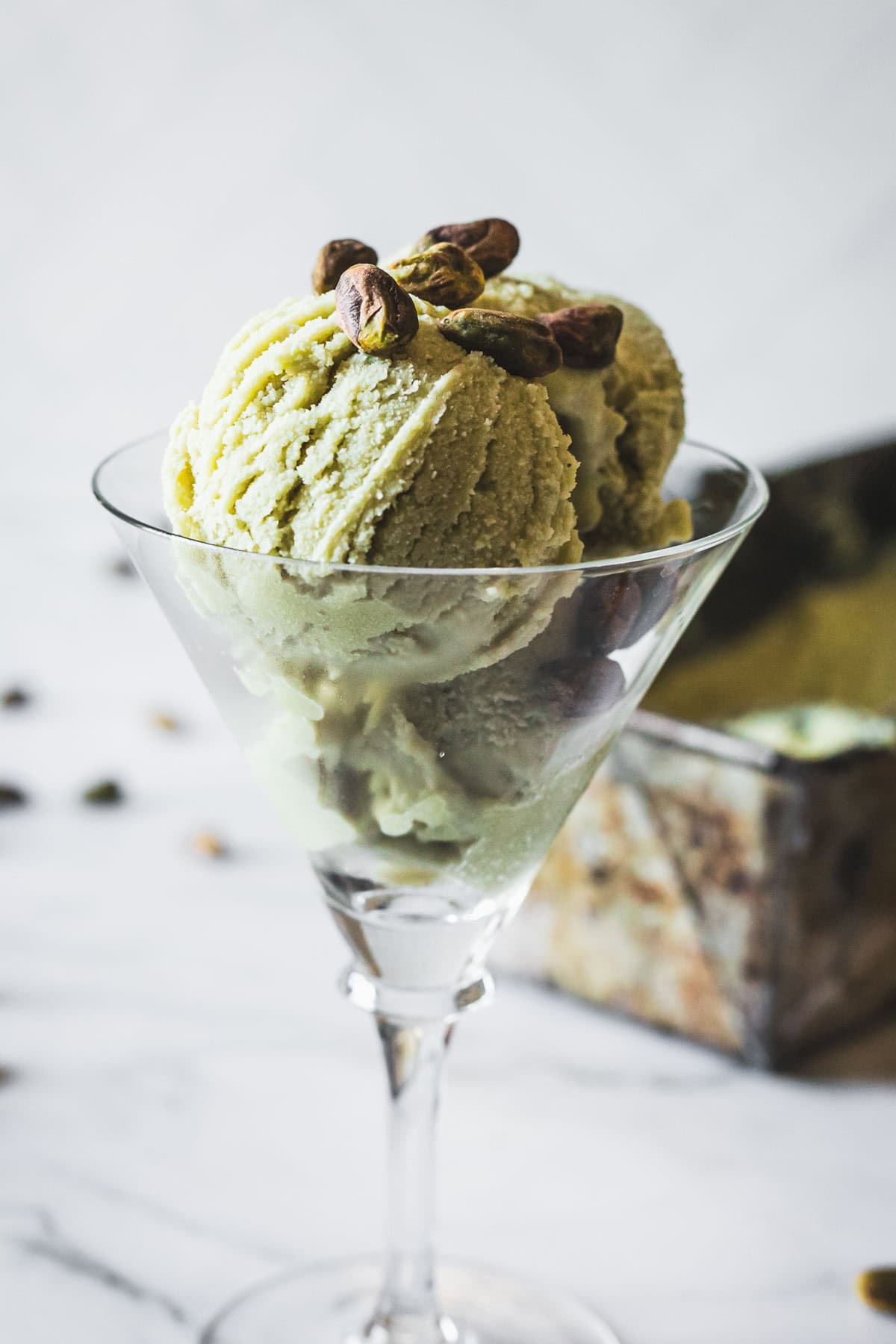 Two scoops of pistachio ice cream in a martini glass, garnished with whole pistachios, with a blurred ice cream container in the background on a white surface.