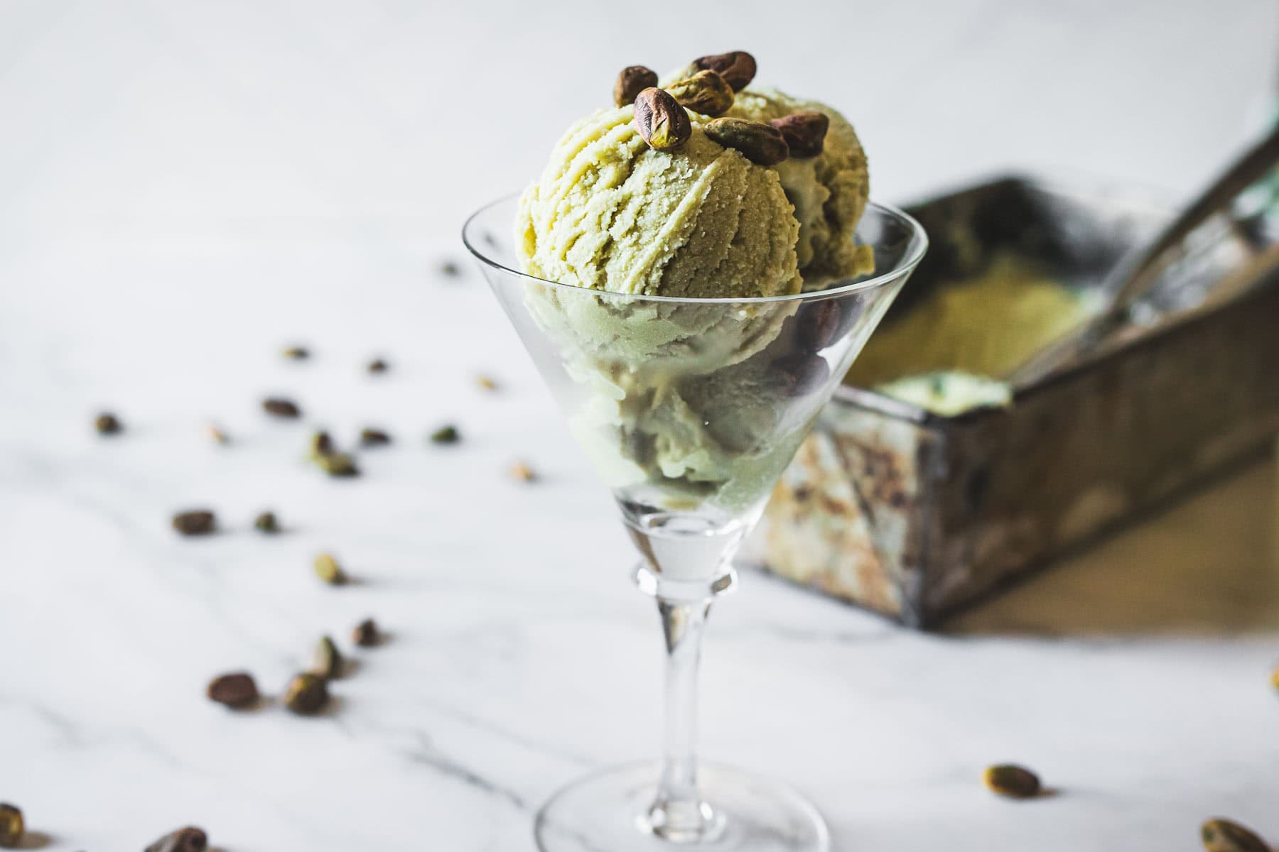Two scoops of Pistachio Ice Cream in a martini glass, garnished with whole pistachios. An ice cream container with a scoop appears in the background, while scattered pistachios rest on the marble surface.