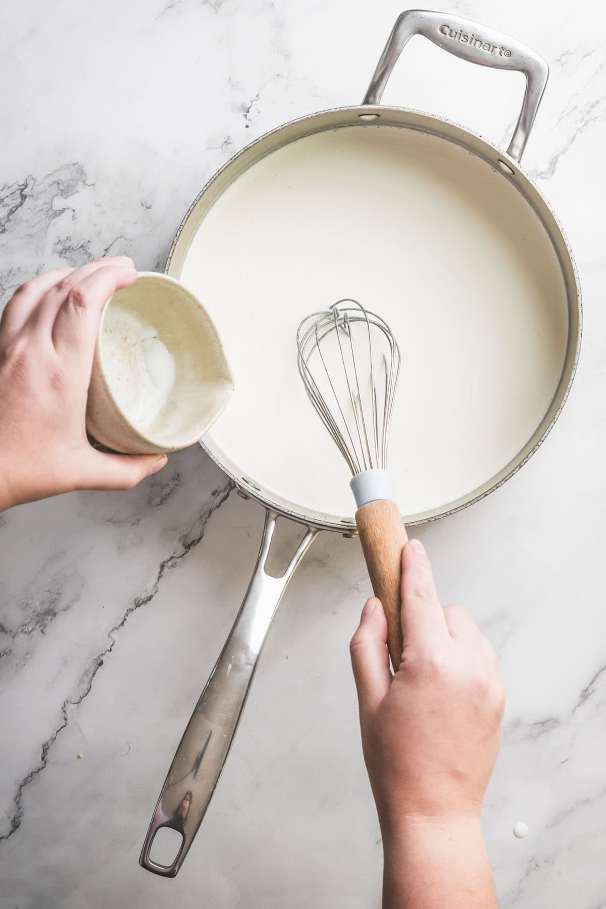 A person holds a whisk in a saucepan filled with a creamy white liquid while pouring a pistachio ice cream mixture from a small cup, on a white marble countertop.