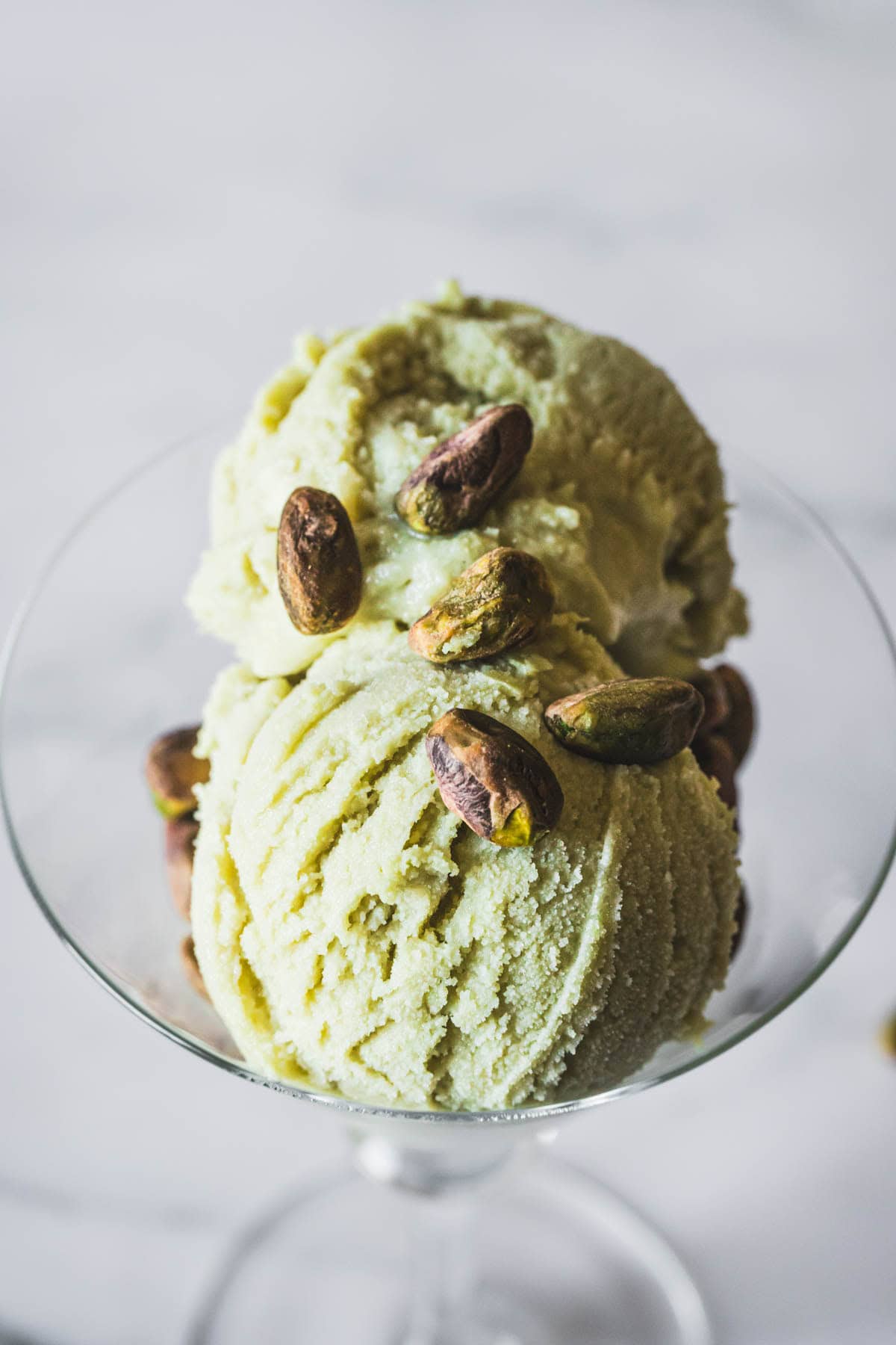Two scoops of pale green pistachio ice cream in a glass dish, topped with several whole pistachio nuts.