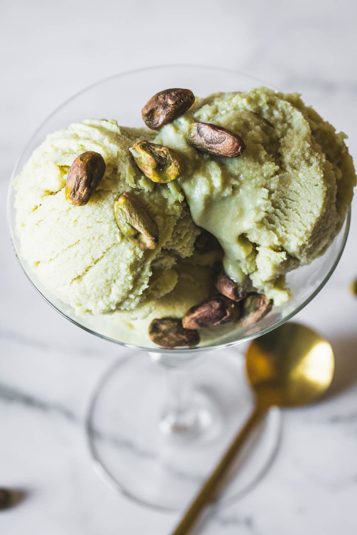 A glass dish filled with two scoops of pistachio ice cream, topped with whole pistachio nuts, sits on a marble surface beside a gold spoon.