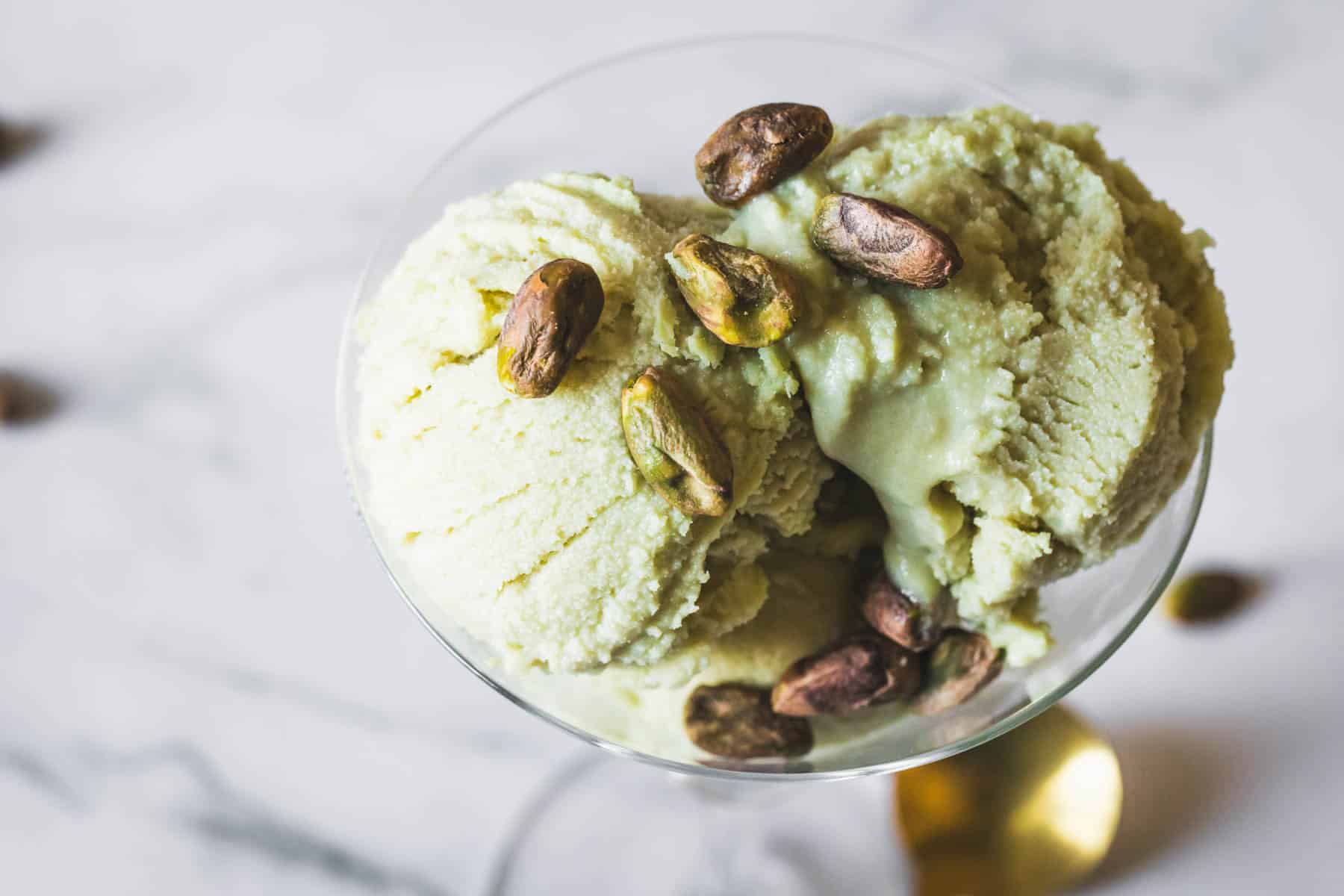 A close-up of three scoops of Pistachio Ice Cream in a glass bowl, garnished with whole pistachio nuts, on a white marble surface.