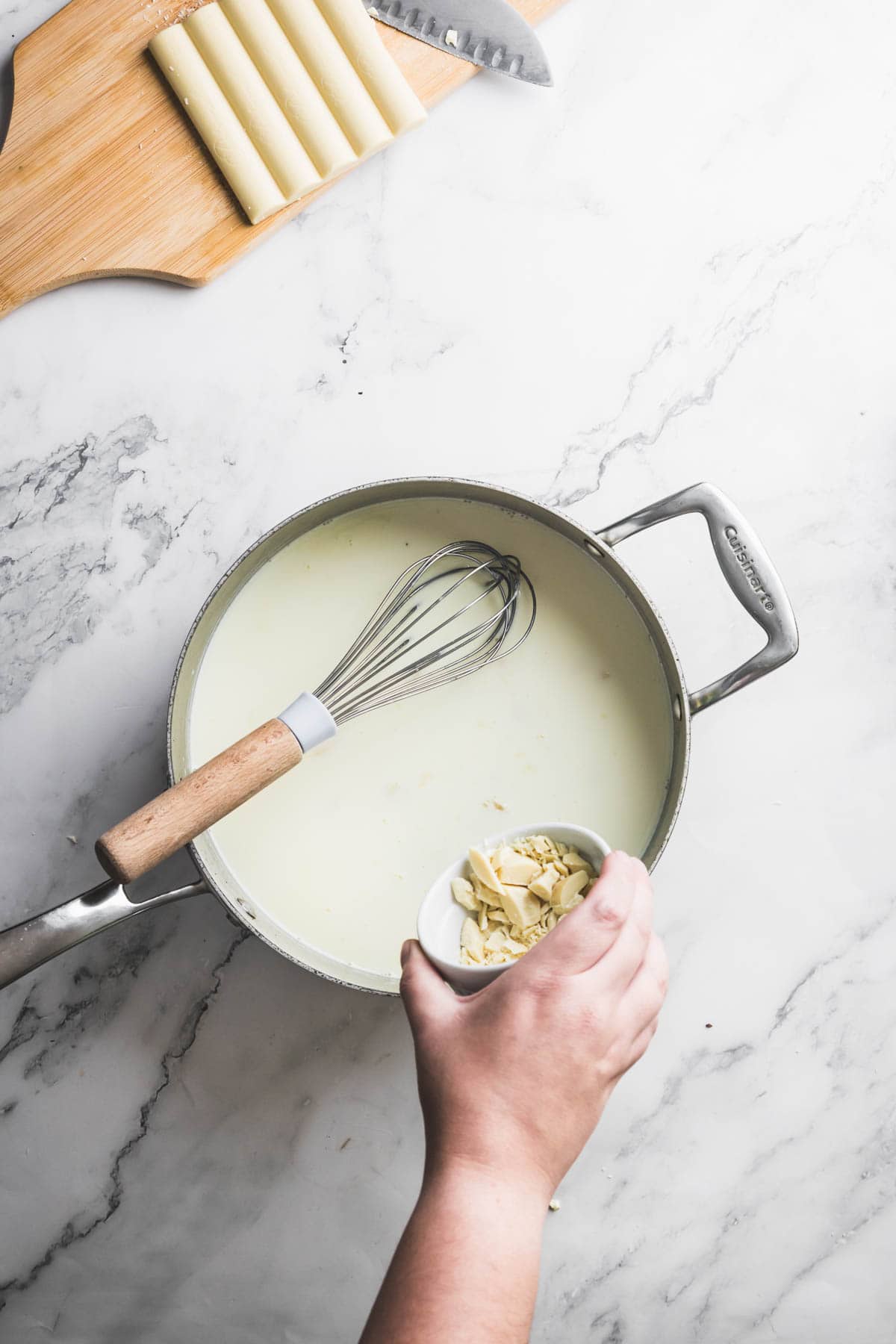 A hand holds a small bowl of white chocolate pieces above a saucepan of light-colored sauce with a whisk inside, preparing a rich base that could be used for Pistachio Ice Cream. In the background, cannelloni tubes and a knife rest on a marble surface.