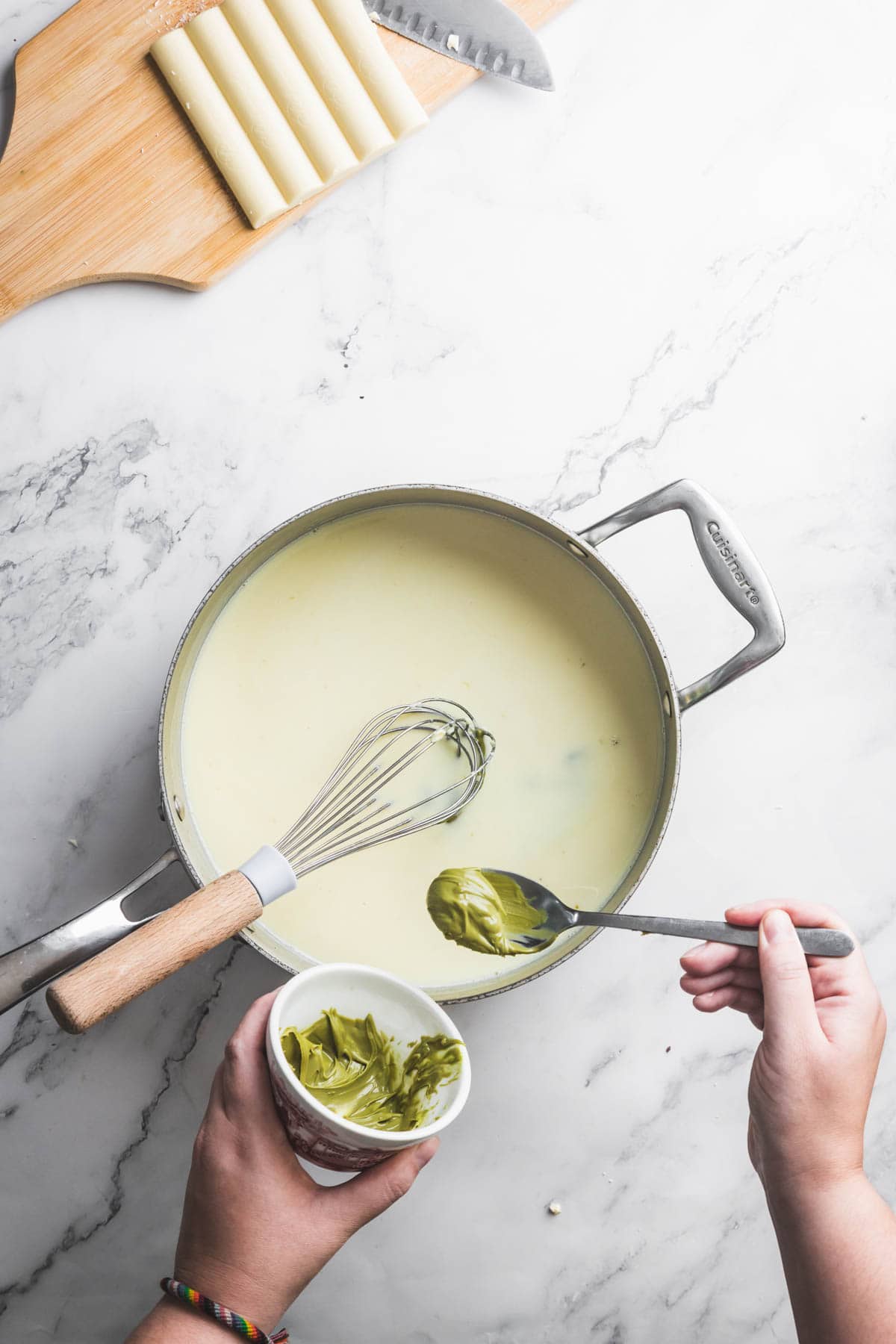 A person holds a spoonful of green pesto over a pot of white sauce on a marble countertop, with a whisk in the pot and a cutting board with pasta tubes and a knife in the background—reminiscent of creamy Pistachio Ice Cream tones.