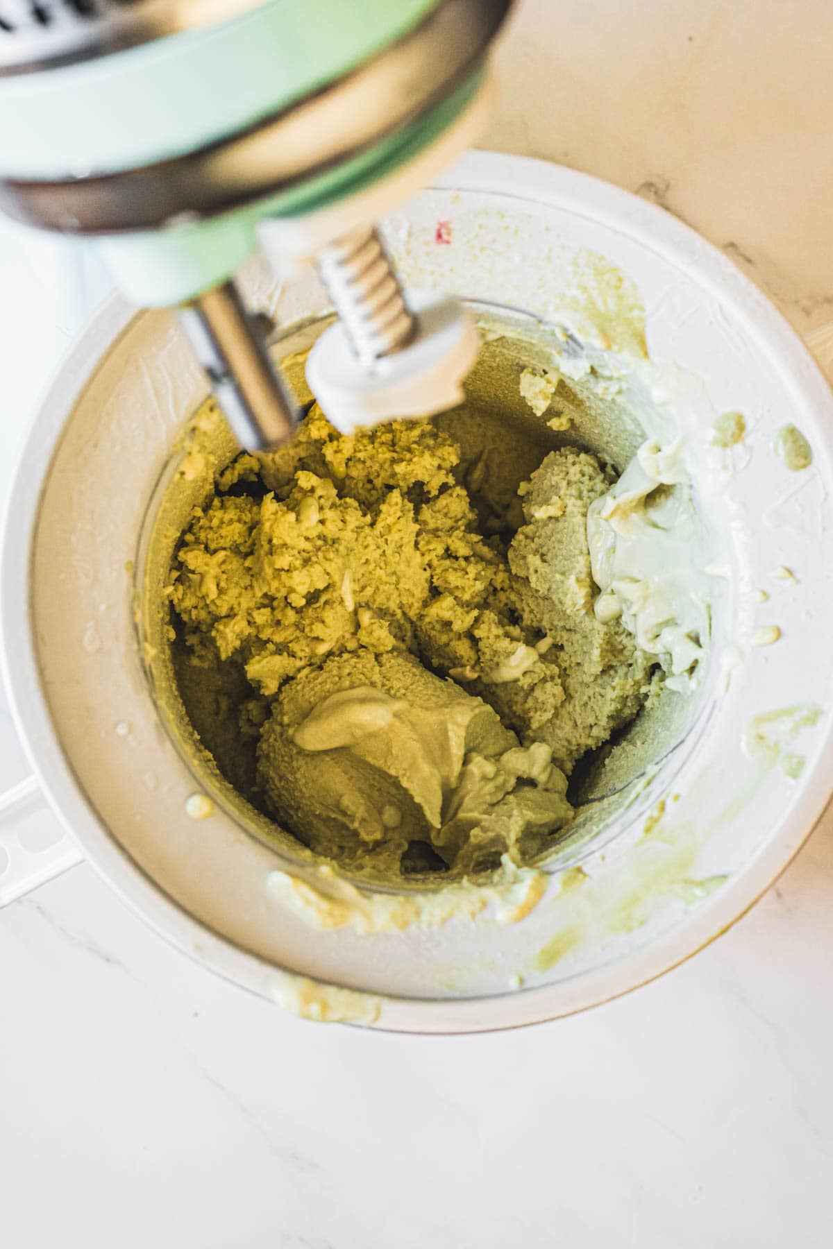 A stand mixer is blending a thick, pale green mixture—likely pistachio ice cream—in a white mixing bowl on a light countertop. Some of the creamy mixture clings to the sides of the bowl.