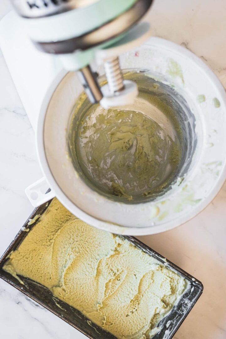 A stand mixer bowl with pistachio ice cream residue inside sits next to a rectangular metal container filled with smooth, freshly churned pistachio ice cream on a marble countertop.