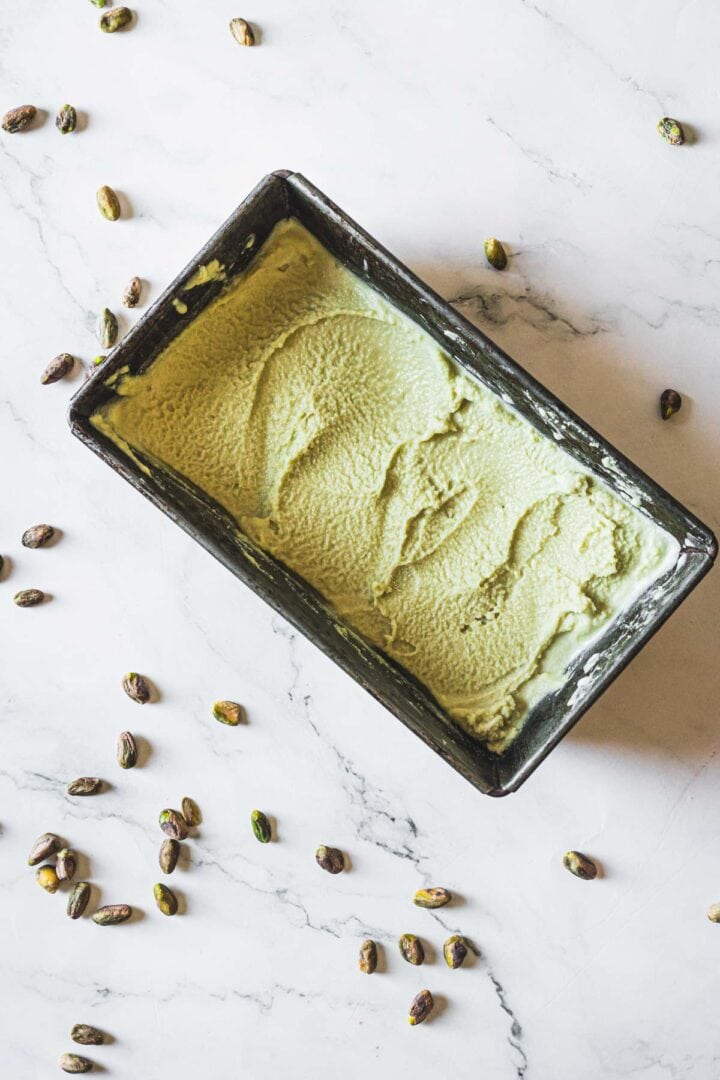 A rectangular metal container filled with creamy Pistachio Ice Cream rests on a white marble surface, surrounded by scattered whole pistachios.