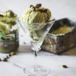 Two scoops of Pistachio Ice Cream served in a martini glass, topped with whole pistachios. A container of pistachio spread and a metal ice cream tray sit in the background on a white surface.