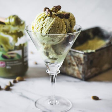 Two scoops of Pistachio Ice Cream served in a martini glass, topped with whole pistachios. A container of pistachio spread and a metal ice cream tray sit in the background on a white surface.