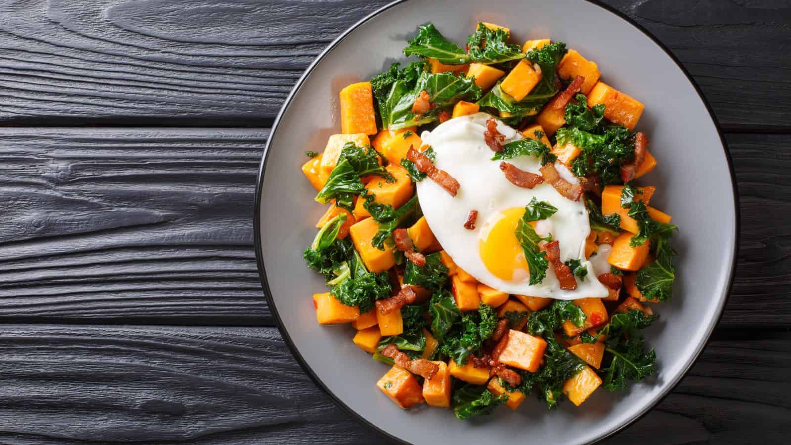 A plate with kale, diced sweet potatoes, bacon pieces, and a sunny-side-up egg on top, served on a gray plate placed on a dark wooden table.