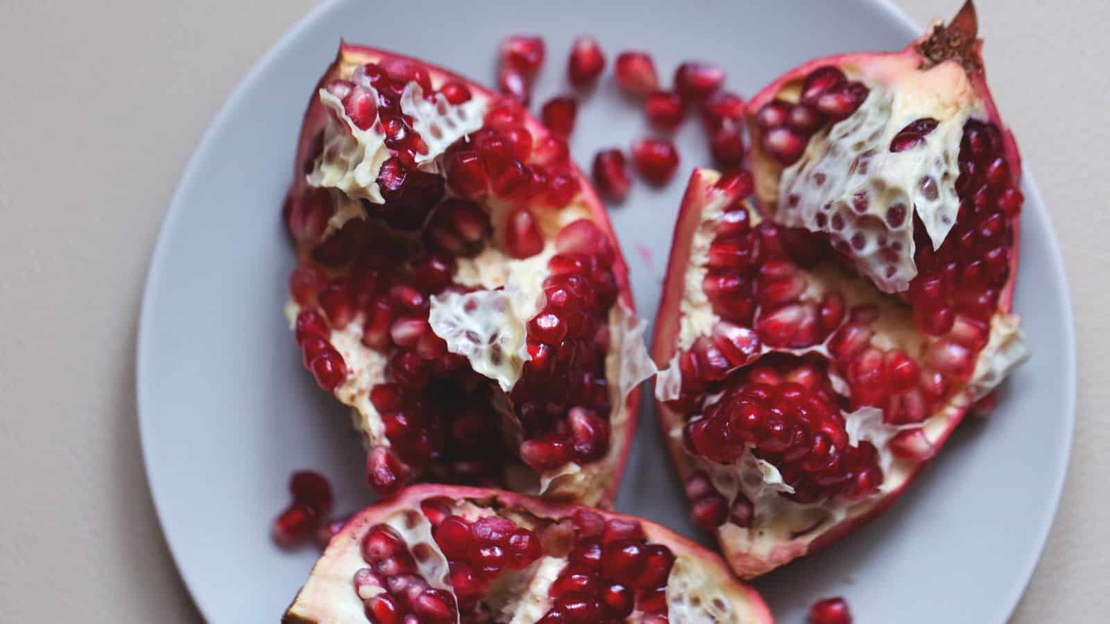 A white plate holds pieces of a split-open pomegranate, showing clusters of red seeds and sections of white pith, with some loose seeds scattered on the plate.