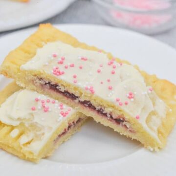 Two rectangular pastry slices with a fruit filling are stacked on a white plate. They are topped with white icing and pink sprinkles. A bowl of pink sprinkles and a gray cloth are in the background.