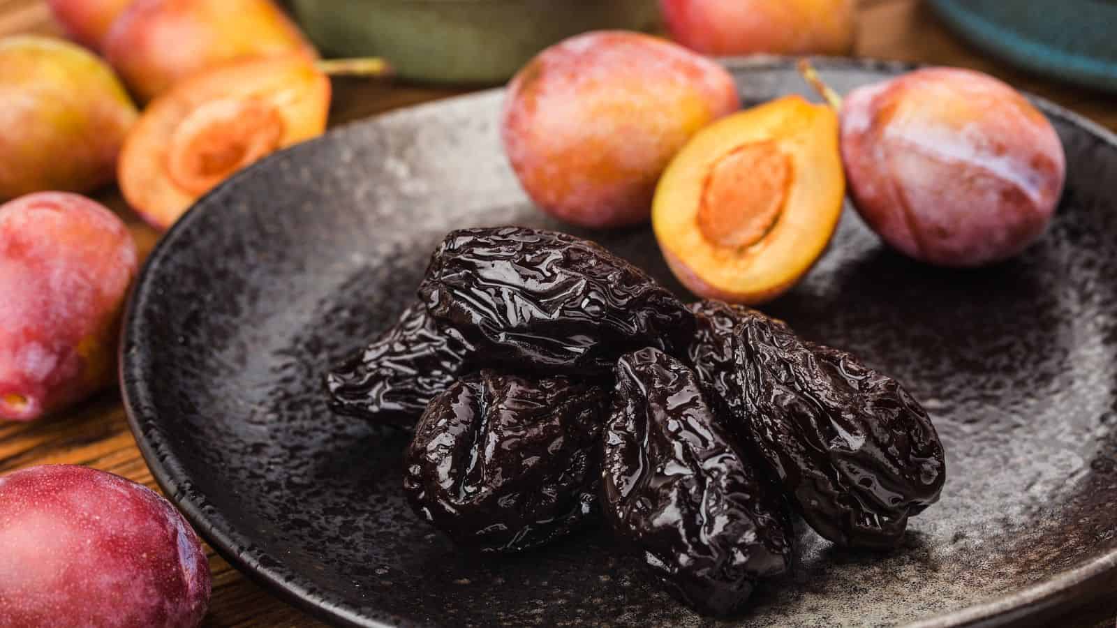 A black plate holds several prunes, with whole and halved fresh plums in the background on a wooden surface.