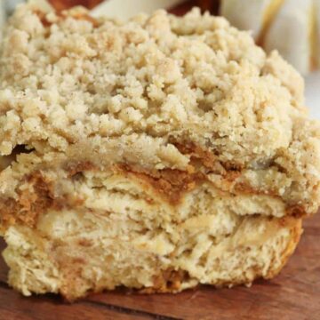 A close-up of a thick slice of crumb cake with a crumbly topping, showing layered, moist cake inside, placed on a wooden cutting board. A small pumpkin is in the blurred background.
