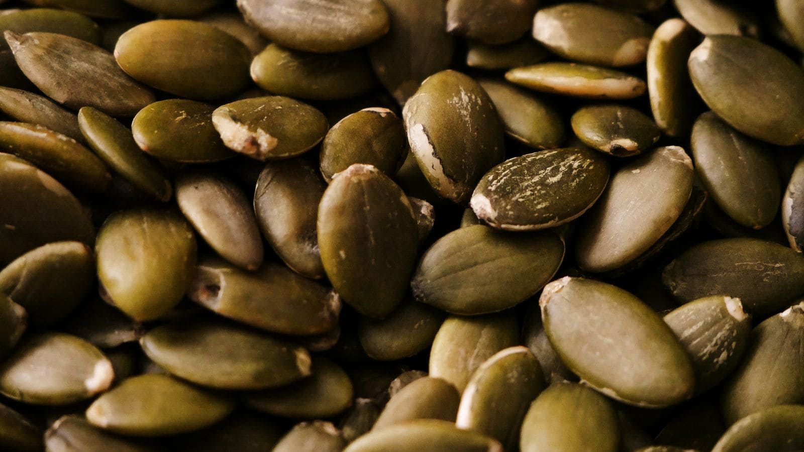 A close-up view of several raw pumpkin seeds with greenish-brown shells, showing their smooth and slightly shiny surfaces.