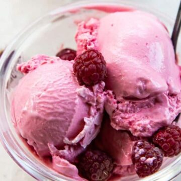 A clear glass bowl contains three scoops of pink raspberry ice cream, garnished with several fresh raspberries. A spoon is placed in the bowl, and a few raspberries are scattered on the white surface nearby.