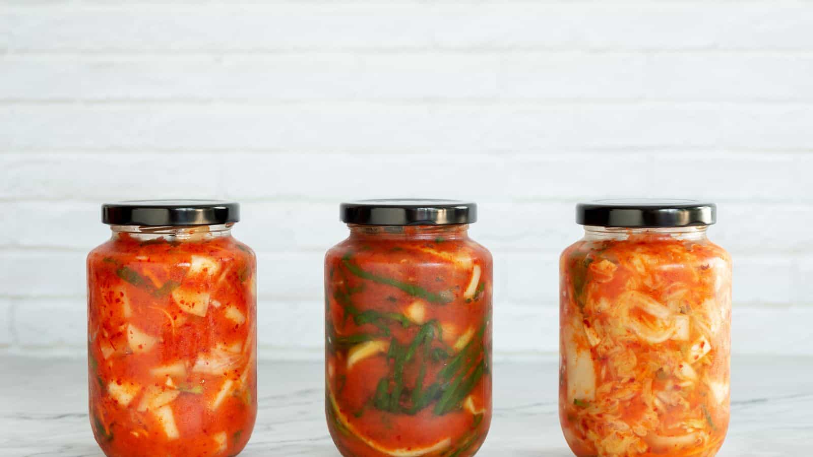 Three glass jars filled with different types of kimchi, each with a black lid, are arranged side by side on a light marble surface in front of a white brick wall.
