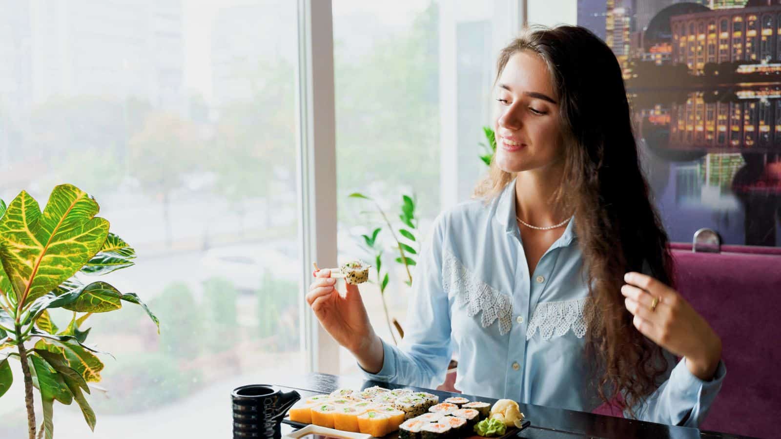 A woman sits at a table by a window, holding a piece of sushi with chopsticks. There is a tray of assorted sushi, a small cup, and a plant on the table. She is smiling and wearing a light blue blouse.