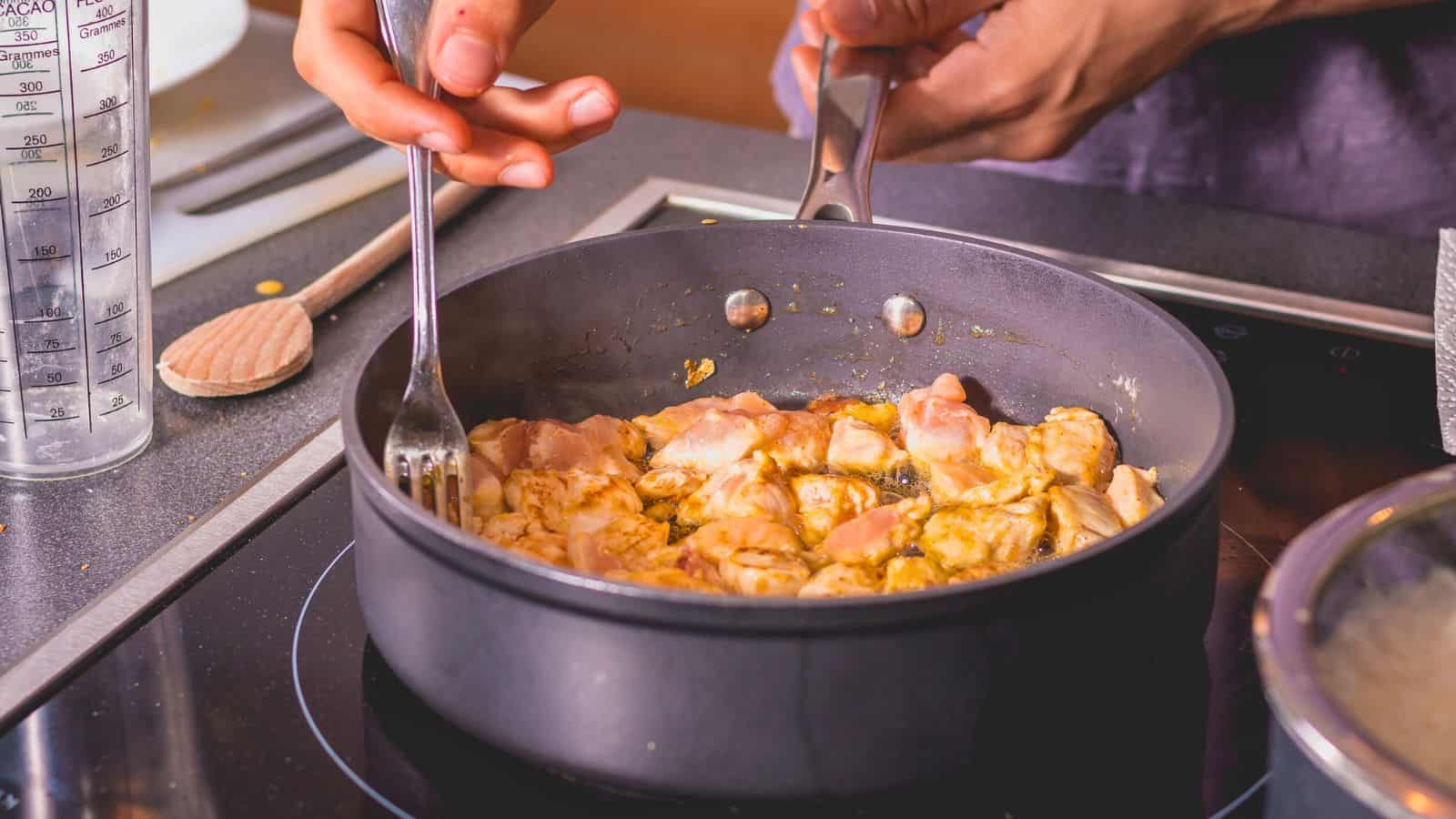 A person cooking diced raw chicken in a frying pan on a stovetop, stirring with a fork while holding the pan handle; a measuring cup and wooden spoon are visible nearby.