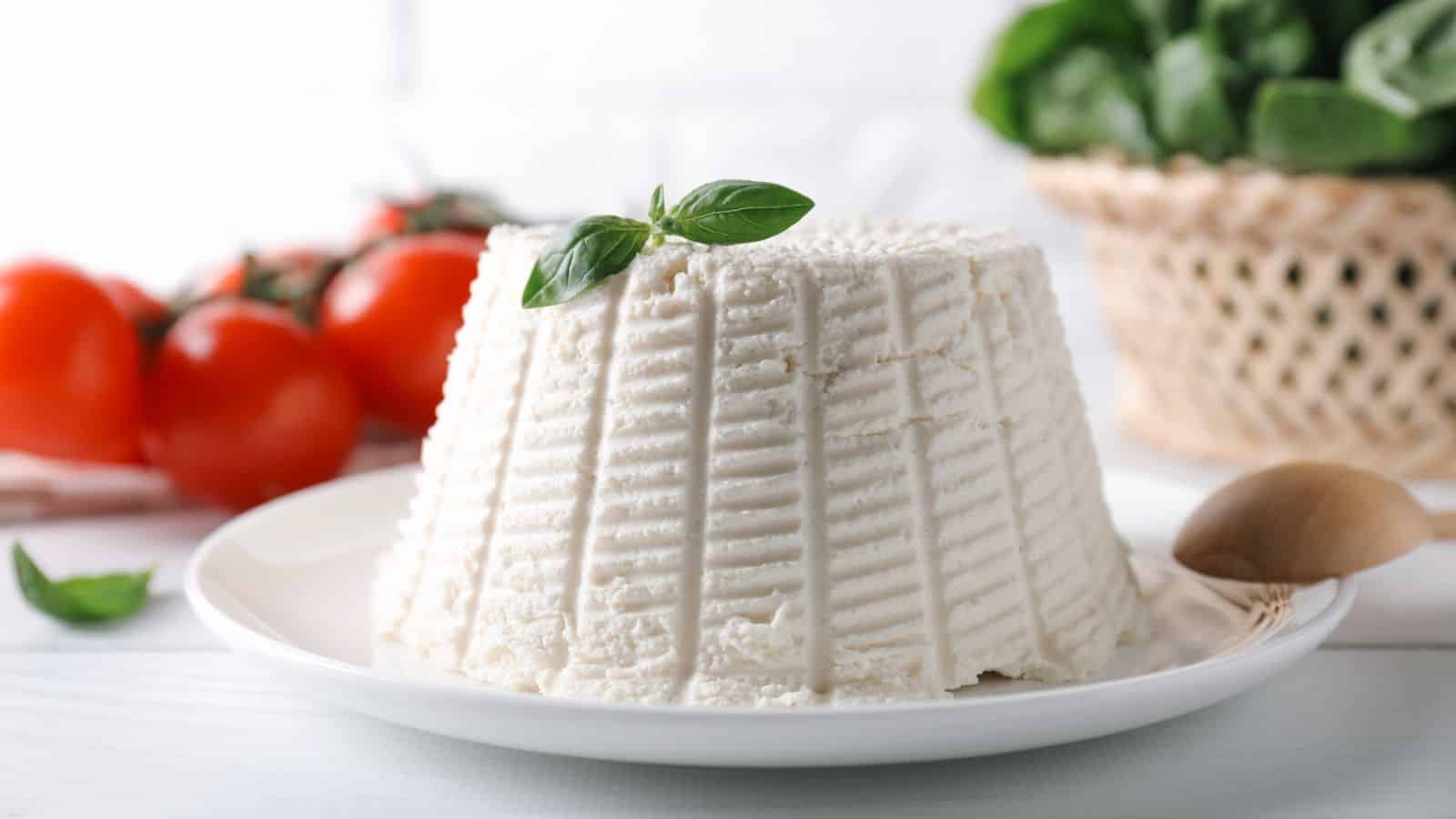 A mound of ricotta cheese sits on a white plate, garnished with a basil leaf. In the background, there are tomatoes, leafy greens in a basket, and a wooden spoon.