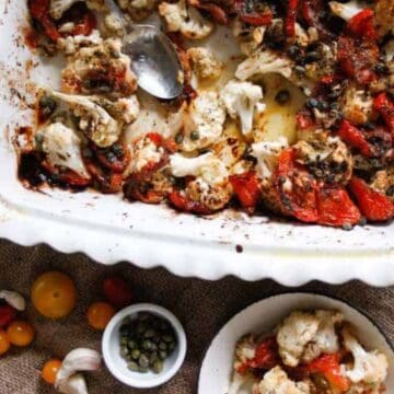 A white baking dish with roasted cauliflower and red peppers sits on a burlap cloth, with a serving spoon, a small plate of the vegetables, cherry tomatoes, garlic cloves, and a small bowl of capers nearby.