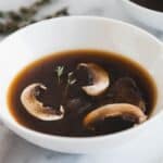 A white bowl filled with clear, brown Roasted Mushroom Broth, containing three mushroom slices and a small sprig of thyme. The bowl sits on a light surface with another bowl partly visible in the background.